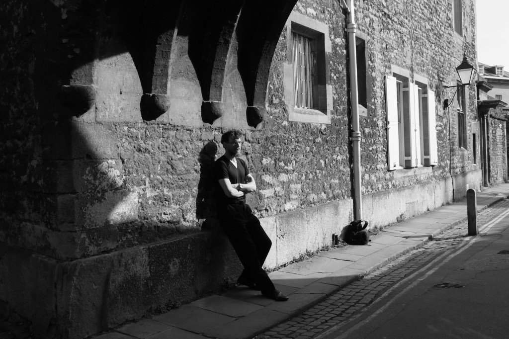 a black and white photo featuring a bearded figure leaning against an old wall under archways, arms folded over the chest and looking contemplative